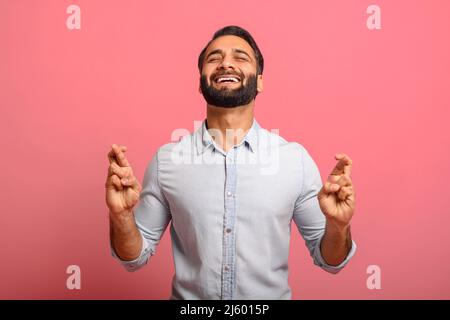 Homme barbu multiracial beau dans un Jean porte-chemise avec les doigts croisés, souhaitant et prière pour le miracle avec les yeux fermés isolé sur fond rose, un Indien montrant le geste pour la chance Banque D'Images
