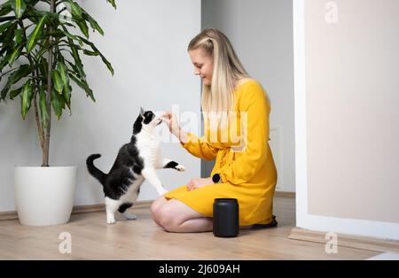 jeune femme blonde avec robe jaune agenouillée sur le sol nourrissant des collations à son chat ayant le pot de traite debout juste à côté d'elle Banque D'Images