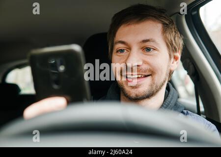 Jeune homme souriant utilisant un smartphone derrière le volant de la voiture. GPS, concept de navigation. Banque D'Images