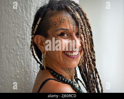 Une jeune femme mexicaine alternative attrayante avec de longues tresses rasta, un piercing facial d'art corporel et des sourires de tatouage positivement optimiste au spectateur. Banque D'Images