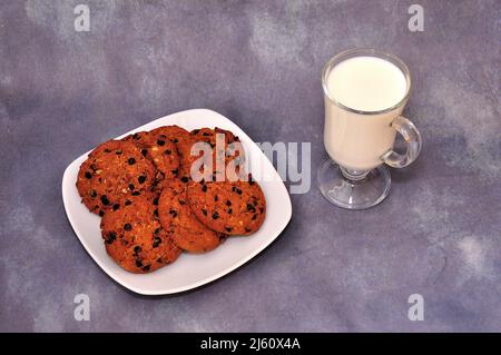 Plusieurs biscuits aux flocons d'avoine et aux pépites de chocolat sur une assiette et un verre de lait chaud sur un fond gris abstrait. Gros plan. Banque D'Images