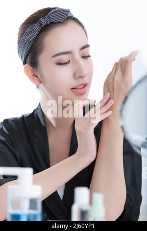 Belle jeune femme chinoise utilisant des produits d'hydratation de beauté devant un miroir de maquillage, sur fond blanc - photo de stock Banque D'Images