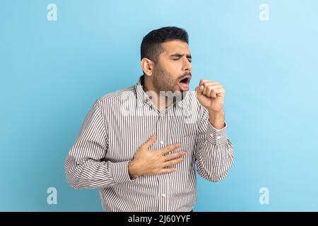 Portrait d'un homme d'affaires barbu malsain toussant, attrape froid, ayant la température élevée, ayant le symptôme de la grippe, portant une chemise rayée. Studio d'intérieur isolé sur fond bleu. Banque D'Images