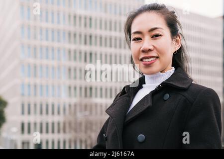 Femme asiatique optimiste en vêtement d'extérieur chic et décontracté souriant gaiement et regardant l'appareil photo sur un arrière-plan flou d'un bâtiment élevé sur la rue de la ville Banque D'Images
