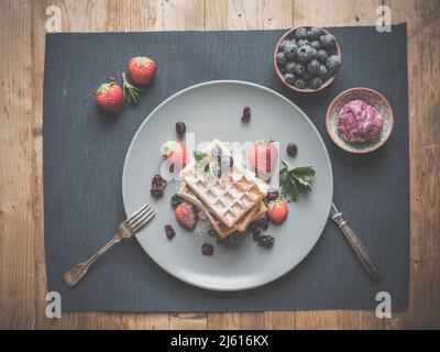 topview de gaufres aux myrtilles sur une assiette bleue et textile décoré avec plus de myrtilles, couverts d'argent, crème glacée à la framboise maison et menthe sur le rusti Banque D'Images