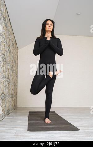 Femme en noir sportswear pratiquant le yoga faisant la variation de vrikshasana exercice, pose d'arbre avec namaste, entraînement debout sur une jambe sur le tapis dans la chambre Banque D'Images