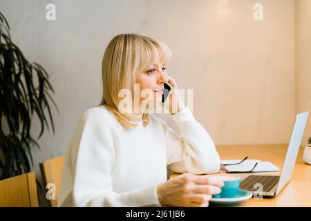 Femme adulte mature assise dans un café avec une tasse de café et travaillant en ligne sur un ordinateur portable. Femme d'affaires parlant sur le téléphone mobile en coworking Banque D'Images