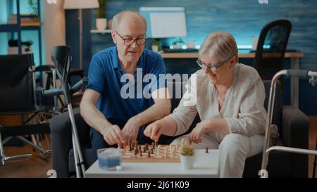 Couple aîné avec cadre de marche et béquilles jouant au jeu d'échecs à bord. Les retraités qui profitent de la concurrence de stratégie avec des pièces d'échecs pour faire cochmate et gagner le match intellectuel. Banque D'Images