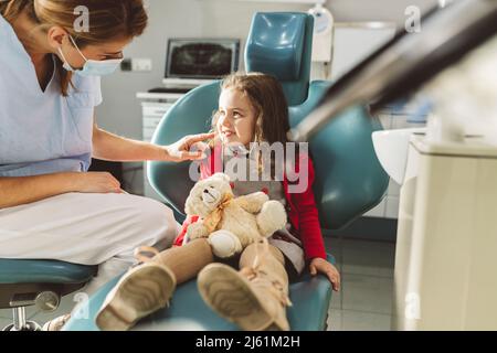 Dentiste dans un masque facial de protection regardant une fille assise avec un jouet bourré dans une clinique dentaire Banque D'Images
