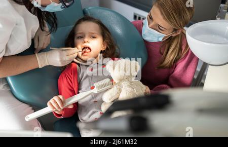 Mère dans le masque facial protecteur regardant la fille examinée par le dentiste à la clinique médicale Banque D'Images