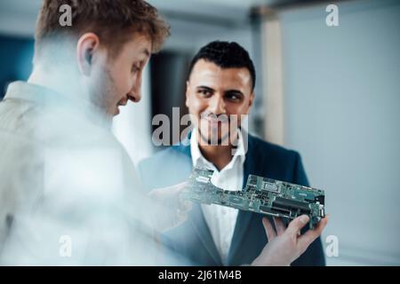 Homme d'affaires souriant regardant un collègue qui analyse la carte de circuit imprimé au bureau Banque D'Images