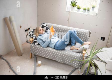 Femme blonde souriante couchée sur un canapé et lisant un livre dans le grenier Banque D'Images