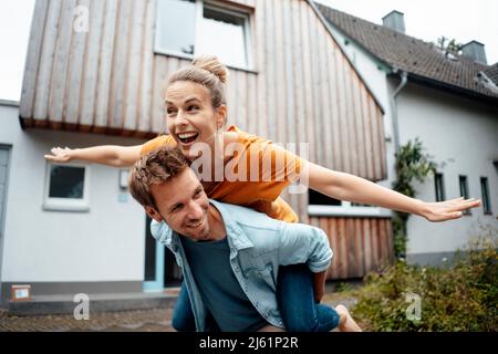 Un homme heureux qui donne une promenade en porcgyback à une femme avec les bras débordés en appréciant à l'arrière-cour Banque D'Images