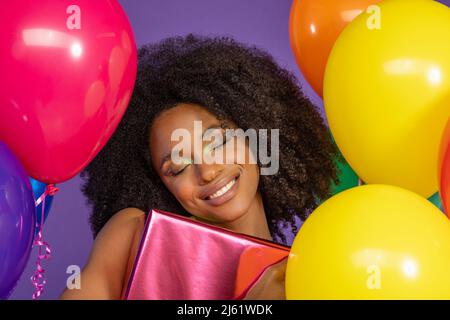 Jeune femme heureuse avec les yeux fermés tenant boîte cadeau debout au milieu de ballons colorés sur fond violet Banque D'Images