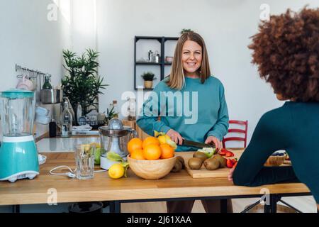 Femme blonde souriante coupant des pommes avec un ami assis à table dans la cuisine Banque D'Images