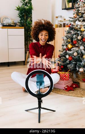 Une jeune femme gaie qui se fait le geste de l'arbre de Noël en se vantant à travers le téléphone portable à la maison Banque D'Images