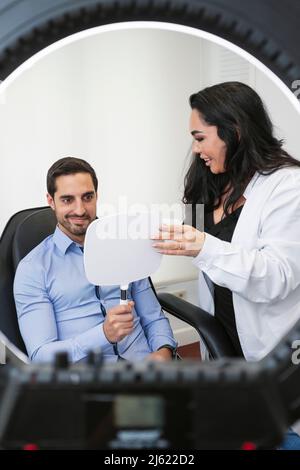 Patient souriant avec un médecin examinant le visage dans le miroir après un traitement de botox à la clinique esthétique Banque D'Images