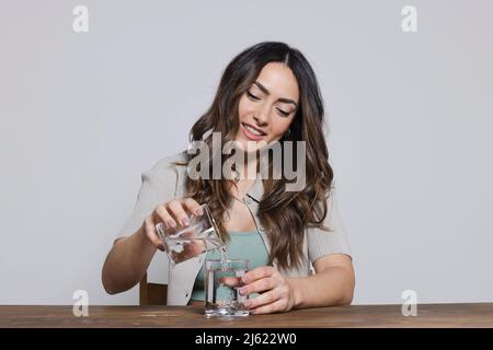 Femme souriante versant de l'eau d'un verre à l'autre assis sur fond gris Banque D'Images