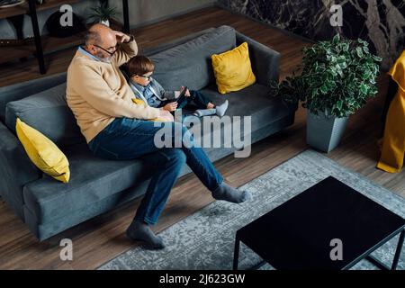 Homme âgé regardant un petit-fils étudiant au moyen d'une tablette PC sur un canapé dans le salon Banque D'Images