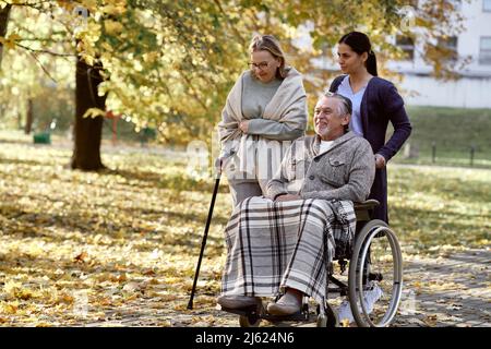 Travailleur de la santé poussant le fauteuil roulant de l'homme handicapé par une femme âgée marchant avec le bâton au parc Banque D'Images