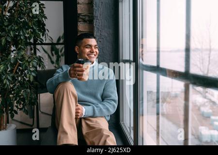 Joyeux jeune homme avec une tasse de café jetable regardant par la fenêtre assis à la maison Banque D'Images