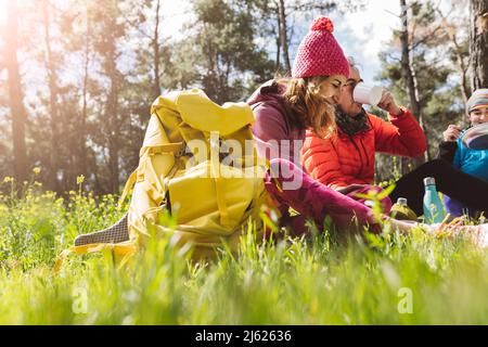 Bonne femme avec sac à dos assis par des amis mangeant et buvant dans la forêt le jour ensoleillé Banque D'Images