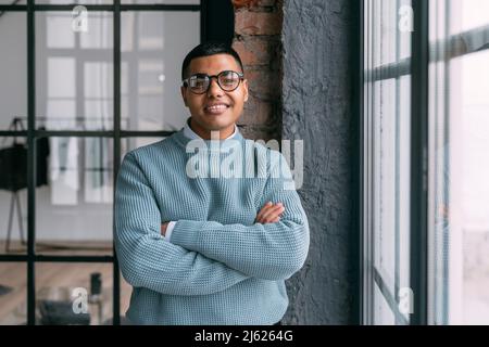 Jeune homme souriant portant des lunettes debout avec les bras croisés par la fenêtre Banque D'Images