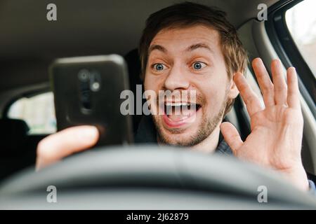 Un jeune homme souriant arrête de conduire la voiture pour répondre à l'appel vidéo sur le smartphone. Banque D'Images