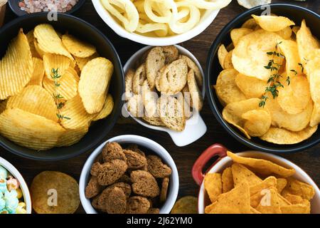 Aliments ou collations malsains. Tous les en-cas classiques de pommes de terre avec des arachides, du pop-corn et des rondelles d'oignon et des bretzels salés dans des assiettes à bol sur fond de bois ancien. Banque D'Images