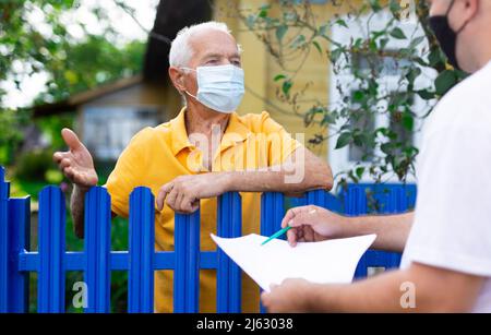 Homme senior en masque de protection communiquant avec le représentant de la compagnie d'assurance tout en se tenant à la clôture de sa maison de campagne Banque D'Images