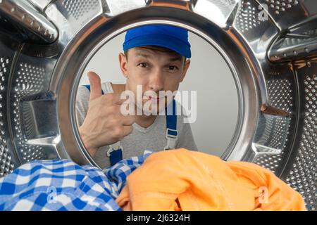 Le réparateur masculin en vêtements spéciaux montre le signe de Thumbs vers le haut, le signe de classe, la photo de l'intérieur du tambour d'une machine à laver. Concept de travail de réparation. Banque D'Images