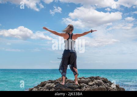 Femme blonde caucasienne perchée au sommet d'un rocher avec ses bras étirés et regardant vers la mer. Sensation de liberté Banque D'Images