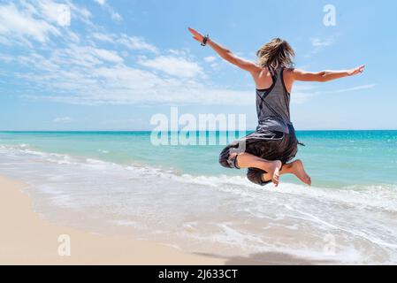 Femme blonde caucasienne sautant pour la joie sur une plage de sable doré et de mer turquoise. Bonheur et plaisir Banque D'Images