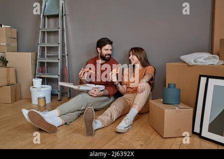 Plan horizontal d'un jeune homme et d'une femme assis ensemble sur le sol en train de manger une pizza dans le salon loft après avoir changé d'appartement Banque D'Images