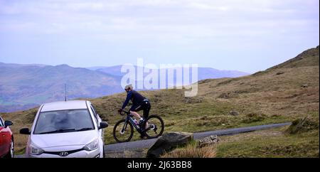 Un cycliste passe devant des voitures garées près du sommet du col Wrynose, Lake District, Cumbria, Angleterre, Royaume-Uni, Îles britanniques. Il y a un nombre limité de places de parking dans la région. Le stationnement est devenu de plus en plus controversé dans la région avec des staycations populaires et tant de automobilistes arrivent maintenant en voiture. « Nous avons l’impression d’être à la pointe de la route », a déclaré Richard Leafe, directeur général du parc national de Lake District (LDNP), en novembre 2021. En 2021, des navettes gratuites ont été offertes dans la vallée de Wasdale et de Cockermouth à Buttermere pour réduire l'impact de l'utilisation de la voiture dans ces vallées populaires. Banque D'Images