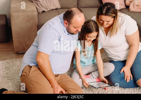 Gros plan d'une famille passant un week-end ensemble à la maison assis sur le tapis regardant la tablette dans les mains de leur fille. Banque D'Images