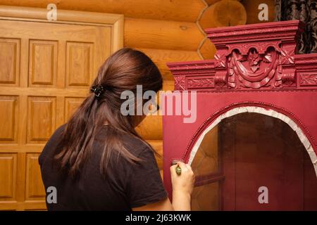 Femme peinture porte de placard en bois avec ornements sculptés avec brosse de couleur rouge. Processus méticuleux de brevelage des meubles. Écologique RE Banque D'Images