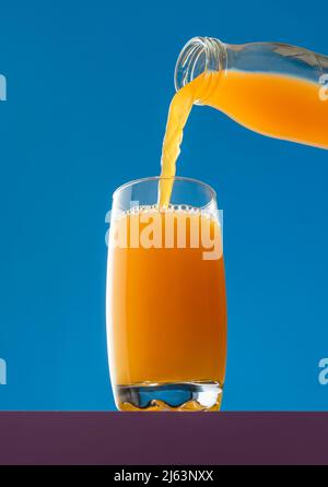 Verser le jus d'orange d'une bouteille dans un verre, isolé sur fond bleu. Vue en angle bas avec un verre de jus d'orange frais. Banque D'Images