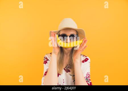 jeune fille blonde avec un chapeau et des lunettes de soleil tenant une banane comme une moustache moyenne gros plan studio tourné orange fond sain concept de style de vie. Photo de haute qualité Banque D'Images