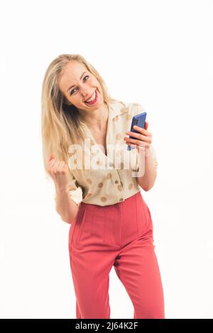 Jolie fille européenne blonde à yeux bleus avec un poing fermé grinning et tenant un téléphone portant une chemise et un pantalon rose moyenne pleine photo blanc fond de copie espace studio tourné. Photo de haute qualité Banque D'Images