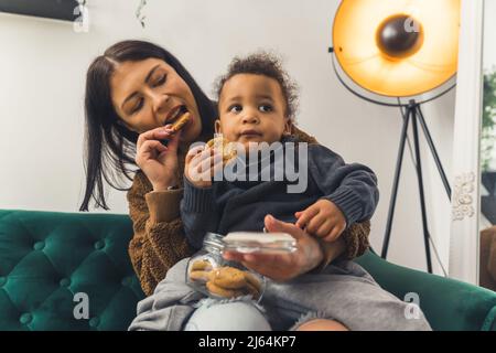 Une jeune mère heureuse et son petit fils biracial assis sur un canapé ensemble et mangeant de savoureux biscuits dans un pot à biscuits. Concept de famille. Photo de haute qualité Banque D'Images