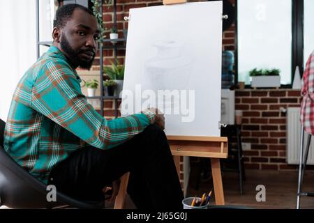 Portrait d'un élève souriant assis devant un vase en toile blanche à l'aide d'un crayon graphique pendant la leçon d'art en studio de créativité. Jeune artiste développant des compétences artistiques pour la croissance personnelle Banque D'Images