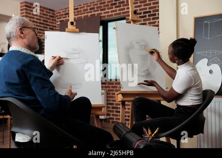 Homme senior enseignant supervisant un jeune étudiant pendant un cours de dessin dans un studio de créativité développant des compétences artistiques. Artiste esquissant un modèle de vase sur toile à l'aide de la technique de dessin. Banque D'Images