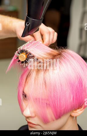 Séchage de cheveux courts roses de jeune femme caucasienne avec un sèche-cheveux noir et une brosse ronde noire par les mains d'un coiffeur de sexe masculin dans un salon de coiffure, gros plan Banque D'Images