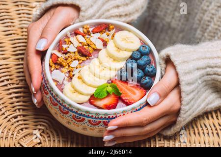 Femme mains tenant des fruits frais açai smoothie bol. Bol à smoothie végétalien sain avec banane, fraise, myrtille et granola. Banque D'Images