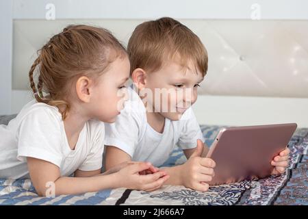 Enfants et gadgets. Un garçon et une fille sont couchés sur le lit, regardant la tablette et souriant Banque D'Images