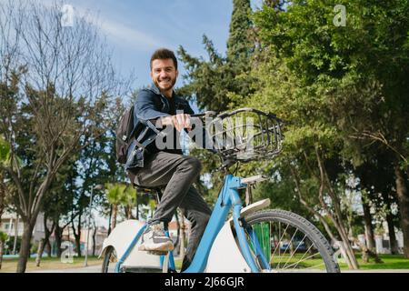 Beau homme regardant la caméra avec sourire pendant une balade en vélo de location dans le parc de la ville, par une journée ensoleillée Banque D'Images