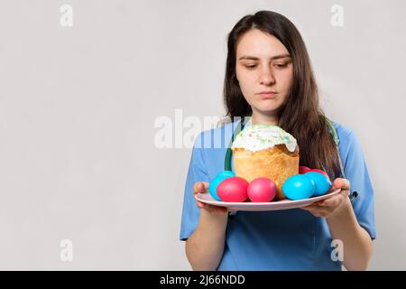 Un professionnel de la santé en uniforme bleu tient un gâteau de Pâques et peint des oeufs sur un fond blanc, un endroit pour le texte. Banque D'Images