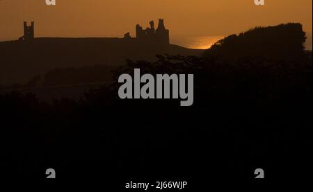 Vue sur le château de Dunstanburgh éloigné en silhouette pendant le coucher de soleil d'automne avec la mer embrassée par le soleil en arrière-plan Banque D'Images