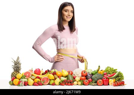 Placez la femme dans des vêtements de sport mesurant sa taille derrière une pile de fruits et de légumes isolés sur fond blanc Banque D'Images